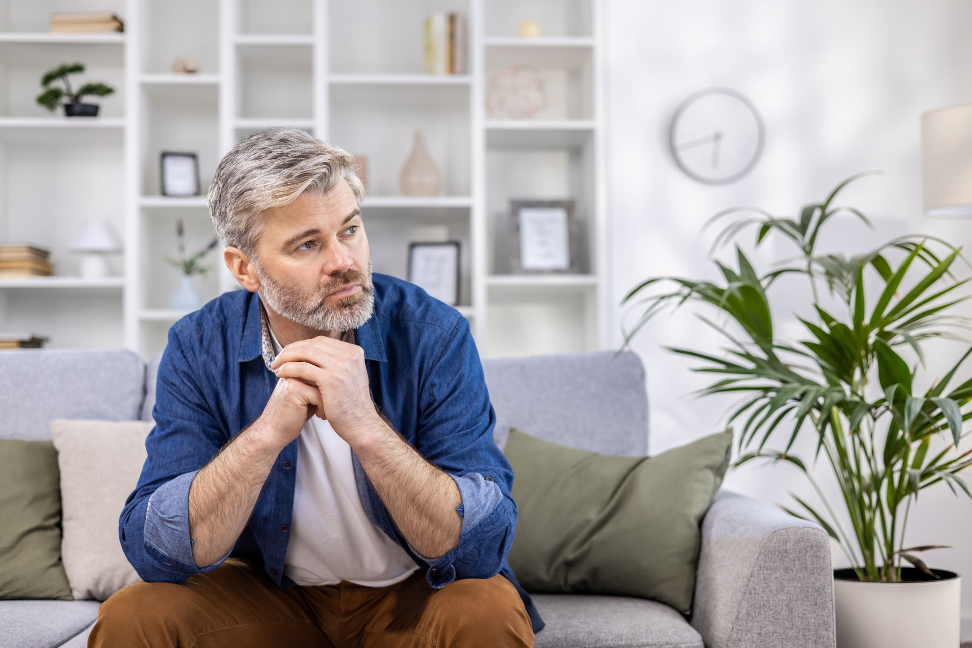 Adult mature man pensive sitting alone at home on sofa close up, gray haired person thinks looks towards window, depressed sad